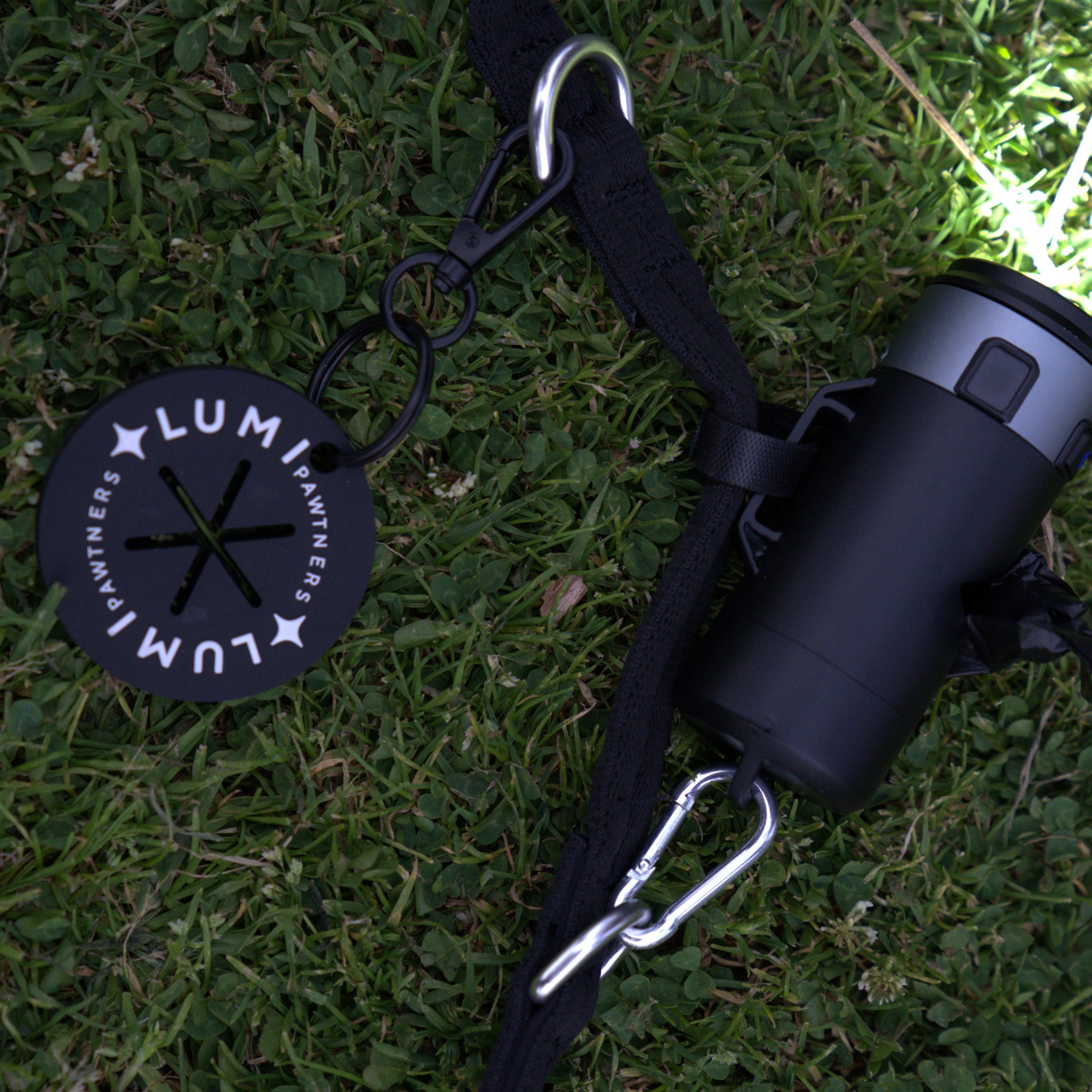 Black waste bag holder with carabiner on grass, featuring 'Lumi' brand logo.